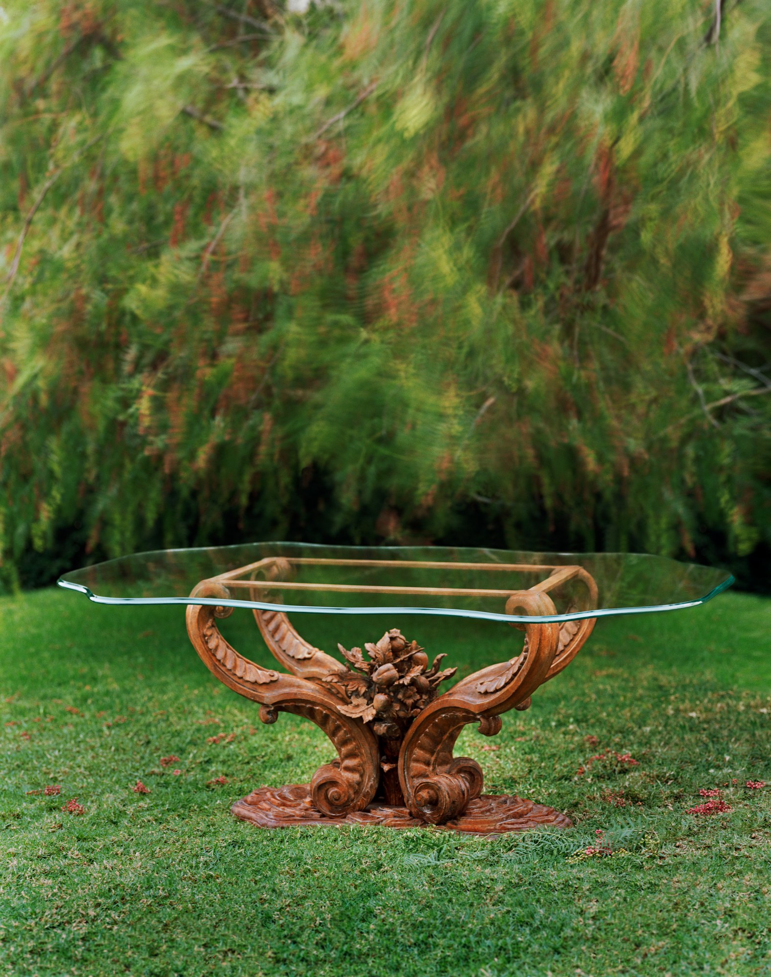 Godeane, a richly carved wooden table base with acanthus scrolls under a shaped glass top, on a lawn