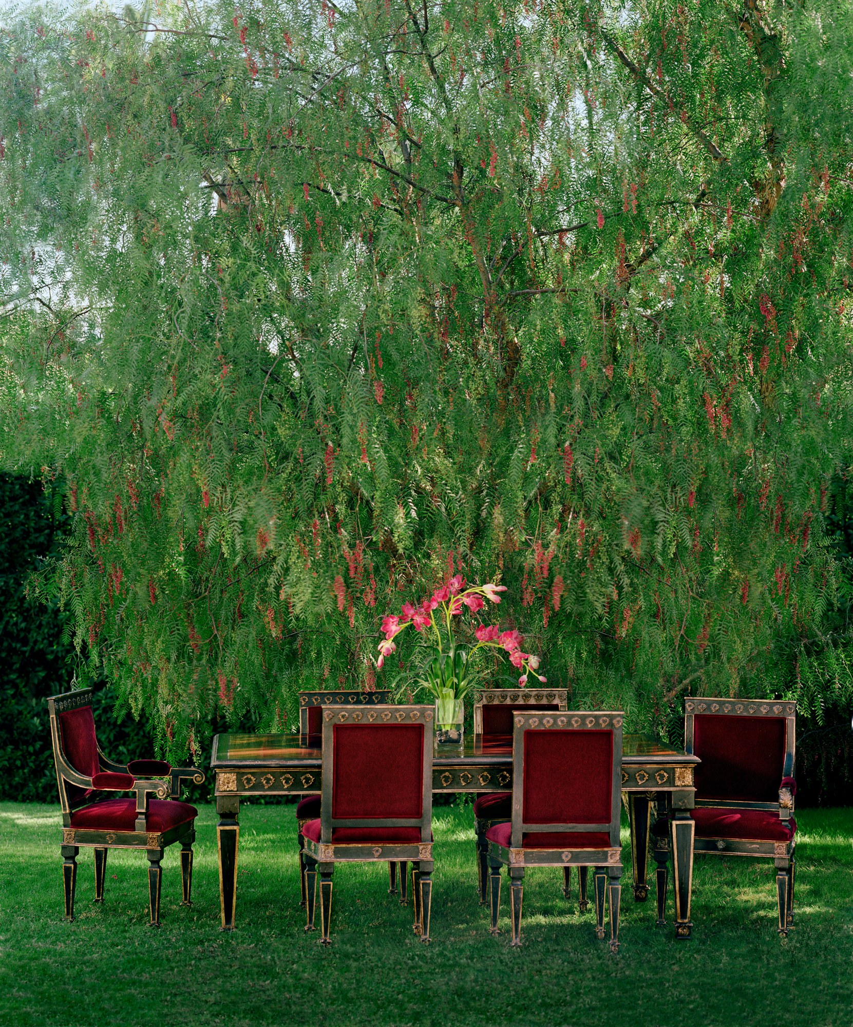Nancee dining table set for a meal on a lawn, surrounded by red upholstered chairs, lilies on the table