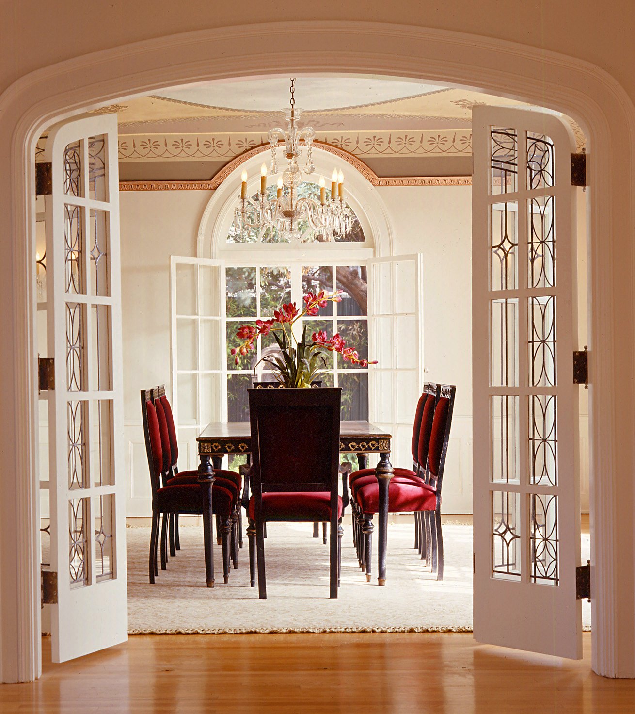 The dining room seen through open leaded French doors, claret chairs beneath a crystal chandelier