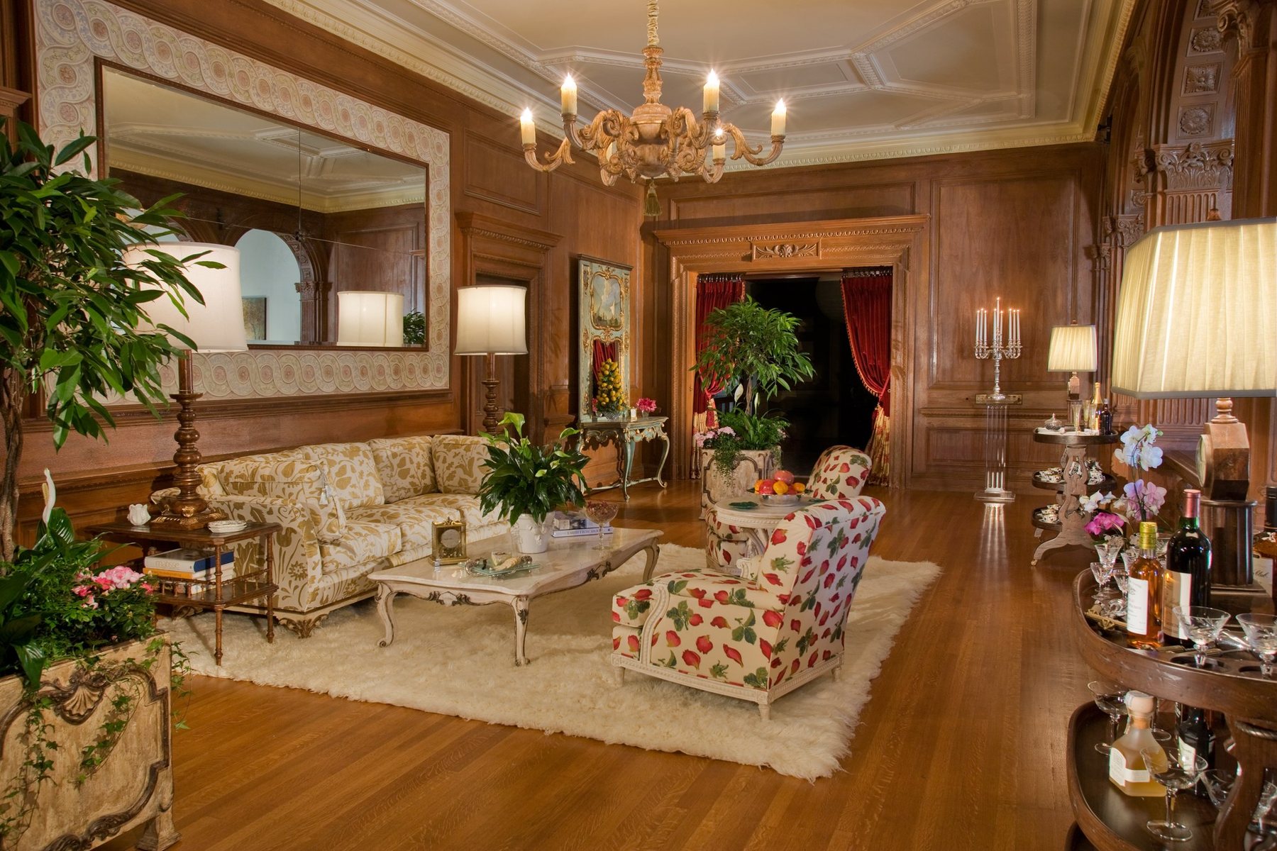 The finished Second Floor Center Hall at Greystone Mansion: oak panelling, a carved ceiling, a crystal-hung chandelier, a gold damask sofa and a strawberry-print chair and ottoman