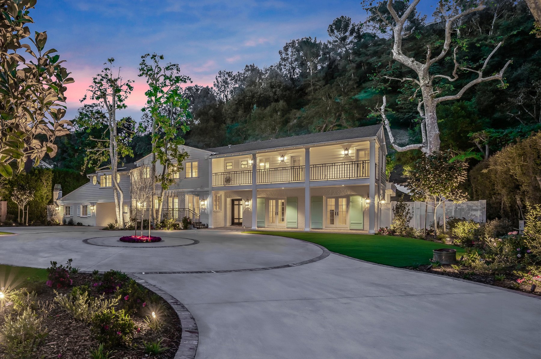 A white two-story Los Angeles house at twilight, its colonnaded porch lit, behind a circular motor court and planted gardens