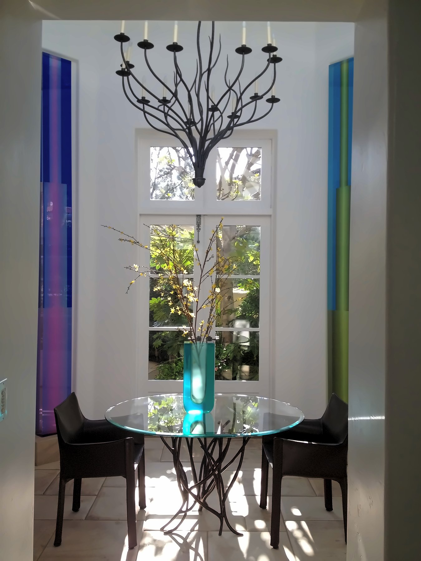 Breakfast nook with an iron branch chandelier over a round glass table, between tall colored glass panels