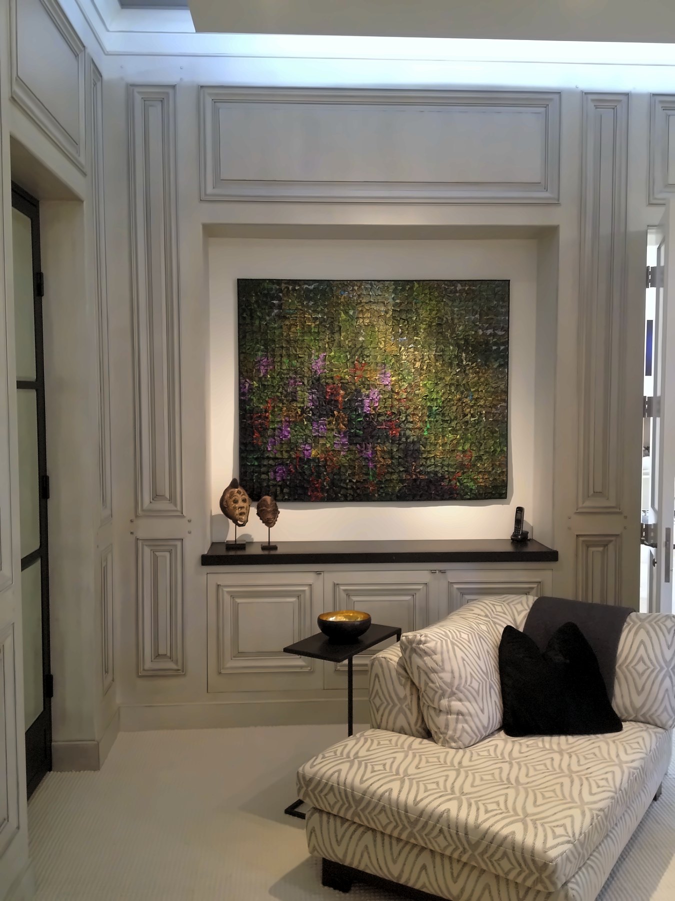 A chaise beneath a large painting set into a lit, paneled niche