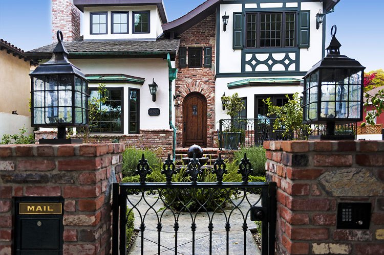 Brick piers with iron lanterns and a wrought-iron garden gate before a Tudor-style house